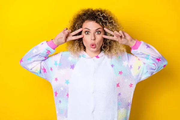 Retrato de atraente surpreendido engraçado menina de cabelos ondulados no pijama mostrando v-sinal perto do olho isolado sobre fundo de cor amarela brilhante — Fotografia de Stock