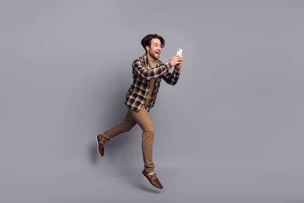 Photo of impressed pretty guy dressed checkered shirt jumping high chatting device isolated grey color background