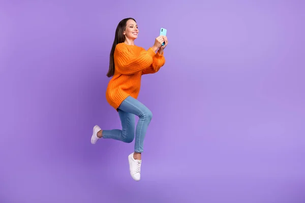 Foto av energiska aktiv dam håll telefonen läsa chat hoppa bära orange pullover isolerad violett färg bakgrund — Stockfoto