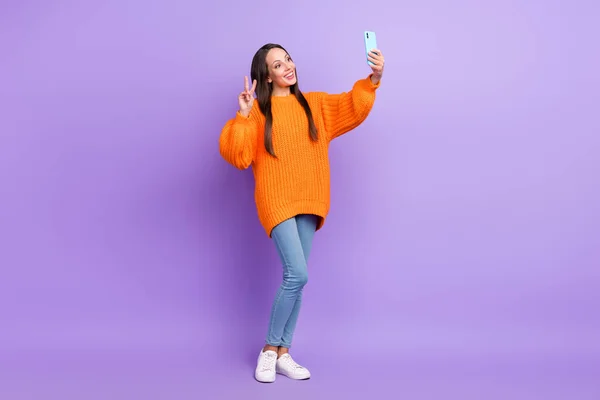 Foto av vänliga söta dam håll telefonen gör selfie show v-tecken bära orange tröja isolerad lila färg bakgrund — Stockfoto