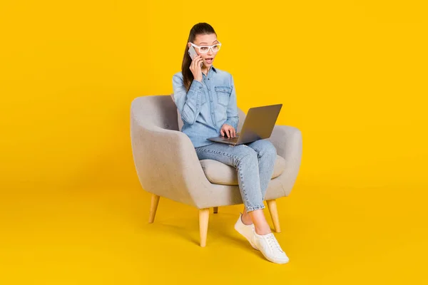 Full length body size woman sitting in chair talking on cellphone using laptop isolated bright yellow color background — стоковое фото