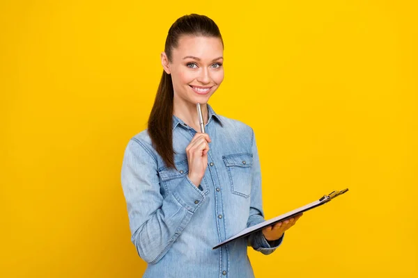 Photo of sweet charming young woman wear denim shirt smiling writing clipboard pen chin isolated yellow color background — Stock Photo, Image