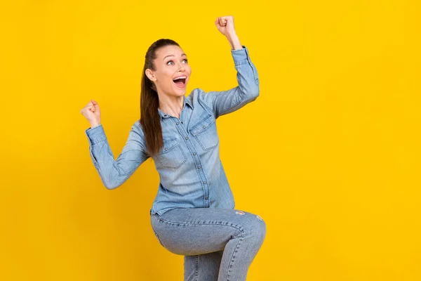 Photo of adorable lucky young lady dressed jeans shirt looking empty space rising fists smiling isolated yellow color background — Φωτογραφία Αρχείου