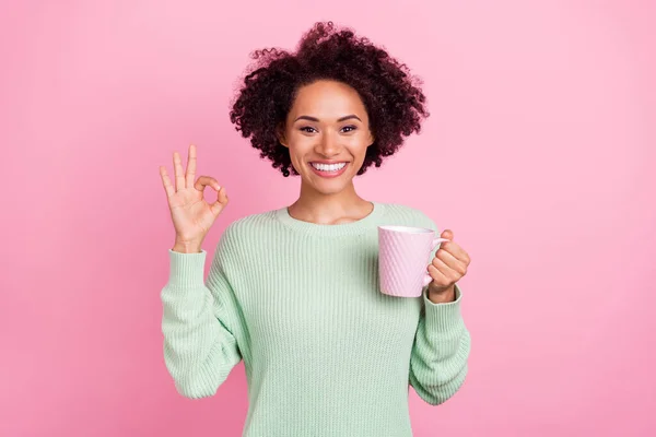 Fénykép a vonzó göndör haj vidám sötét bőr hölgy tart tea bögre megjelenítése okey szimbólum elszigetelt rózsaszín színű háttér — Stock Fotó