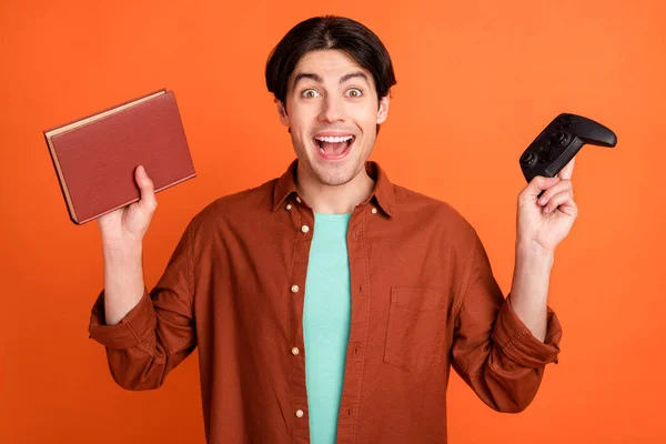 Portrait of young smiling excited scream shout hold playstation and book alternative choice isolated on orange color background — Stockfoto