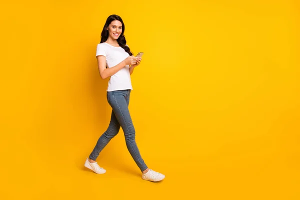 Full längd kroppsstorlek attraktiv glad vågigt hår flicka som går med hjälp av enheten gadget isolerad över ljusa gula färg bakgrund — Stockfoto