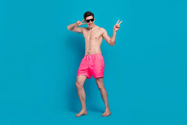 Foto de loco despreocupado clubber hombre mostrar v-signo de desgaste sunglass traje de baño pantalones cortos aislados de color azul de fondo — Foto de Stock