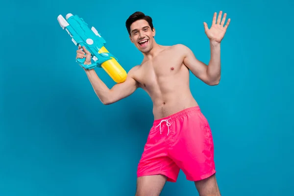 Foto de funky loco juguetón chico mantenga bomba pistola onda mano desgaste traje de baño pantalones cortos aislados de color azul fondo — Foto de Stock
