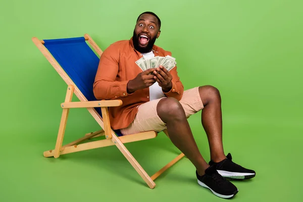 Full size foto van onder de indruk millennial brunet guy houden geld dragen shirt shorts schoenen geïsoleerd op groene achtergrond — Stockfoto