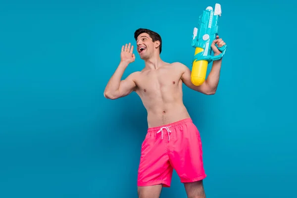 Foto de adorable joven divertido torso desnudo sosteniendo pistola de agua haciendo anuncio espacio vacío sonriendo aislado color azul fondo — Foto de Stock