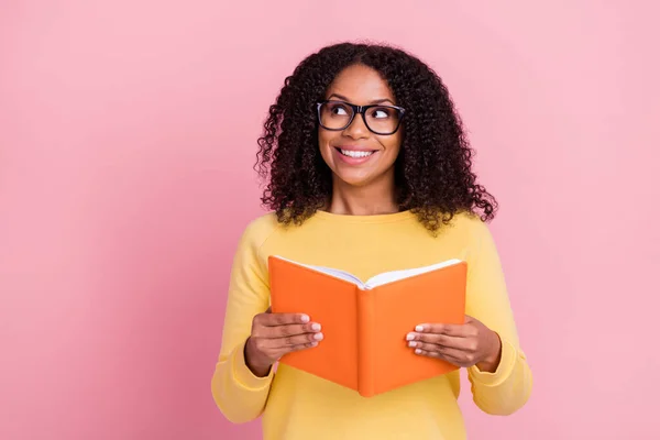 Foto de niña de piel oscura soñadora excitada vestida con gafas de jersey amarillas sosteniendo libro buscando espacio vacío aislado color rosa fondo — Foto de Stock