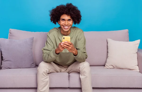 Foto de hombre joven utilizar el teléfono celular como blogger popular suscríbete sit divan aislado sobre fondo de color azul —  Fotos de Stock