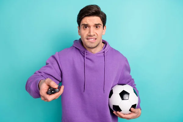 Фото нервного расстроенного молодого парня, одетого в фиолетовую толстовку, держащего футбольный мяч, переключающий телеканалы — стоковое фото