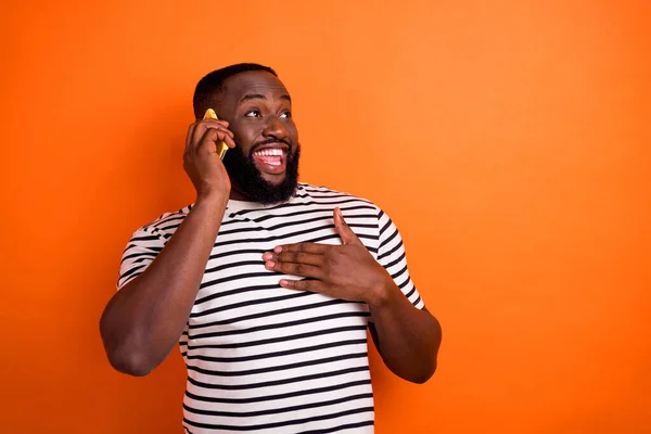 Foto av ung afrikansk man förvånad ser tom utrymme prata mobiltelefon hand beröring bröstet tacksam isolerad över orange färg bakgrund — Stockfoto