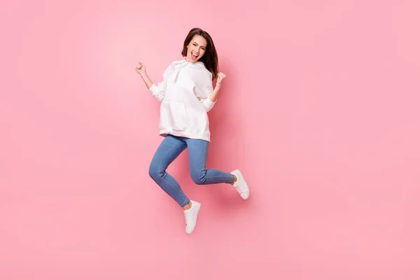 Foto de mujer joven afortunada impresionada vestida con sudadera blanca levantando puños saltando alto sonriendo aislado color rosa fondo —  Fotos de Stock