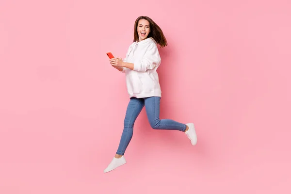 Pleine photo du corps de mignon jeune femme brune sauter avec téléphone porter un jean à capuche blanc isolé sur fond de couleur rose — Photo