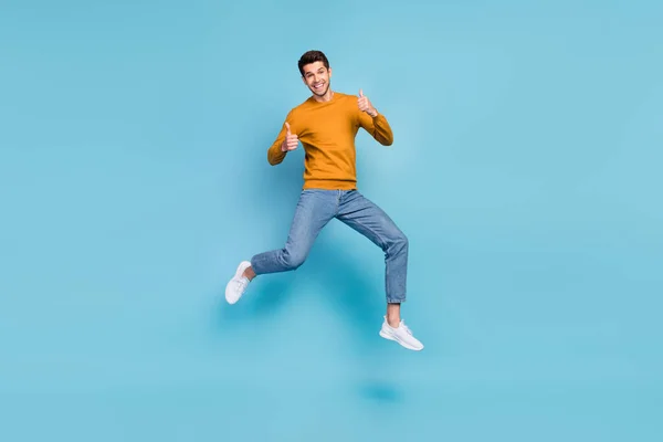 Foto av funky söt ung man bära gul tröja leende hoppa högt visar tummen upp isolerad blå färg bakgrund — Stockfoto
