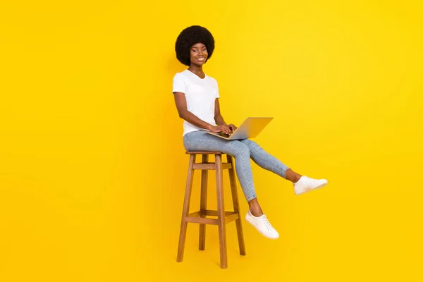 Foto von ziemlich süß dunkle Haut Dame tragen weißes T-Shirt sitzen Barstuhl Typisierung modernes Gerät lächelt isoliert gelbe Farbe Hintergrund — Stockfoto