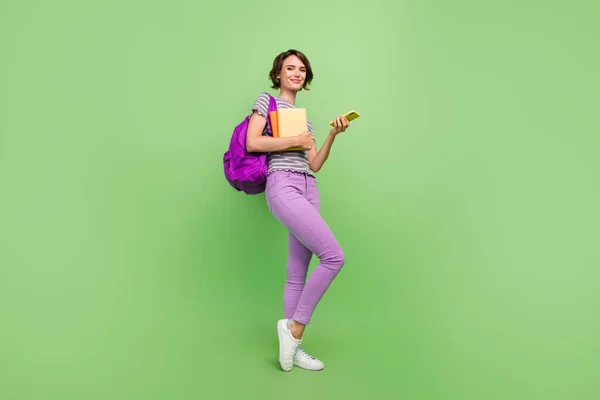 Foto de divertida mujer bastante joven vestida a rayas t-shirt mochila copybooks dispositivo moderno sonriendo aislado color verde fondo — Foto de Stock