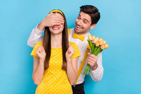 Foto van knappe man arm dekking ogen aantrekkelijk dame houden verse tulpen geïsoleerd op blauwe kleur achtergrond — Stockfoto