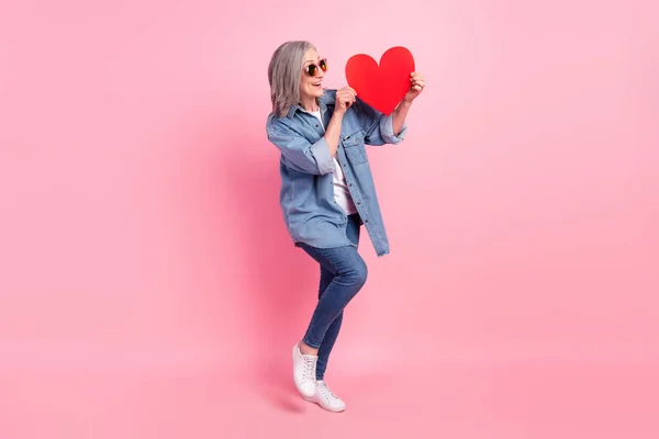 Full tělo fotografie flirtující starší dáma držet srdce nosit modré košile džíny tenisky izolované na růžové barevné pozadí — Stock fotografie