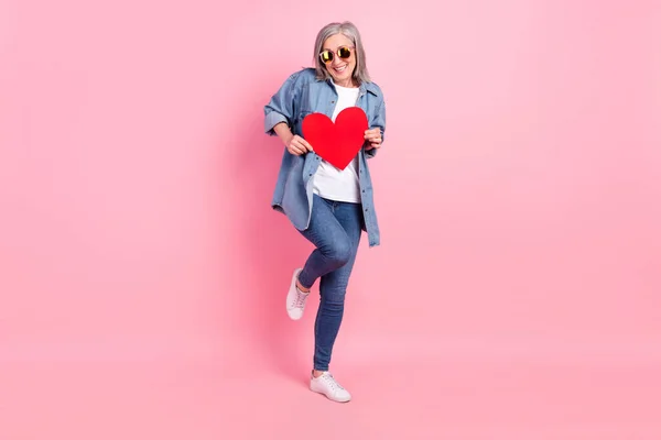Full size φωτογραφία του γλυκού ηλικιωμένη κυρία δείχνουν την καρδιά φορούν μπλε πουκάμισο τζιν αθλητικά παπούτσια απομονώνονται σε ροζ φόντο χρώμα — Φωτογραφία Αρχείου