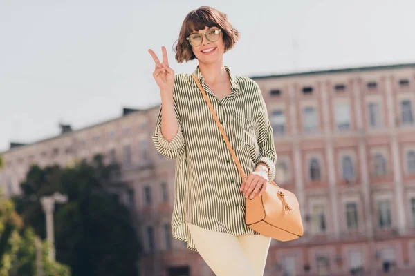 Zdjęcie pozytywne słodkie piękne lady pokazać v-znak promienie uśmiech nosić okulary torba paski koszulka miejskie miasto na zewnątrz — Zdjęcie stockowe