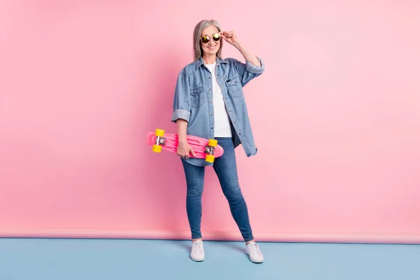 Foto em tamanho completo de cool old lady touch eyewear hold skate desgaste jeans camisa isolada no fundo rosa — Fotografia de Stock
