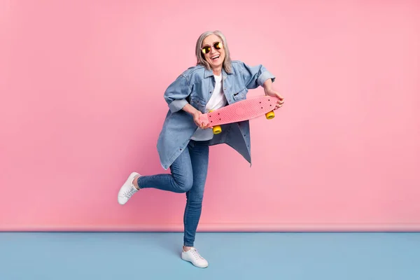 Πλήρης φωτογραφία του σώματος της τρελής ηλικιωμένης κυρίας κρατήσει skate πάει να φορέσει γυαλιά τζιν πουκάμισο απομονώνονται σε ροζ φόντο — Φωτογραφία Αρχείου
