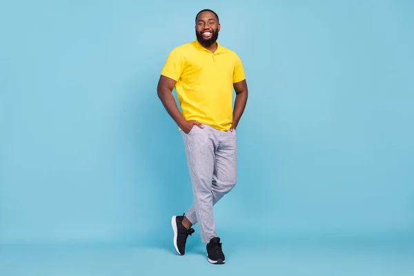 Foto av glada glänsande mörk hud kille klädd gul t-shirt leende armar fickor isolerad blå färg bakgrund — Stockfoto