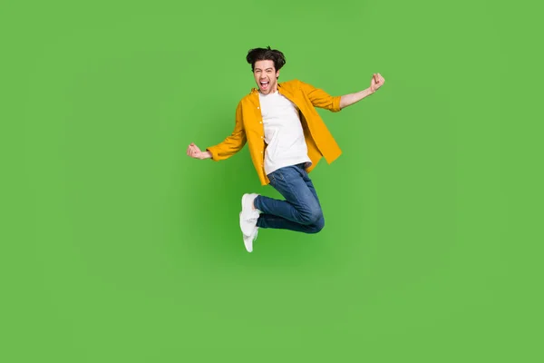 Foto di carino impressionato giovane ragazzo vestito camicia gialla saltando alti pugni in aumento sorridente isolato colore verde sfondo — Foto Stock