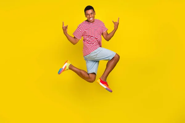 Full length foto di ragazzo salto fare cornuto punk segno usura a strisce t-shirt pantaloncini calzature isolato su sfondo di colore giallo — Foto Stock