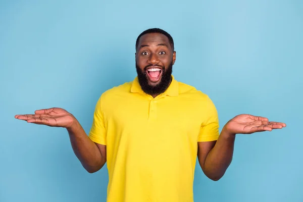 Foto von süß aufgeregt dunkle Haut Mann tragen gelbes T-Shirt lächelnd vergleichen Arme leeren Raum isoliert blaue Farbe Hintergrund — Stockfoto