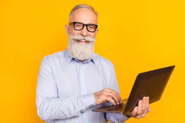 Foto de atraente homem velho positivo segurar laptop desgaste óculos programador isolado no fundo cor amarela brilho — Fotografia de Stock