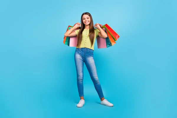 Фото положительной маленькой леди держать розничные пакеты, позируя износа зеленый топ джинсы обувь изолированный синий цвет фона — стоковое фото