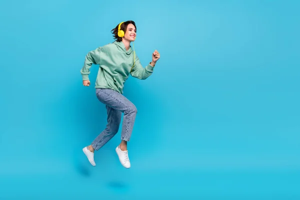 Full längd profil foto av söt tusenårig brunett dam springa bära hörlurar pullover jeans skor isolerad på blå bakgrund — Stockfoto