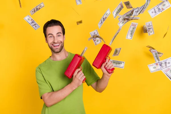 Foto de la barba alegre millennial chico con pistola de dinero llevar camiseta verde aislado sobre fondo de color amarillo — Foto de Stock