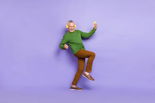 Фото взволнованного пенсионера одетого пуловер танцует поднимая кулаки слушать музыку гаджет изолирован фиолетовый цвет фона — стоковое фото