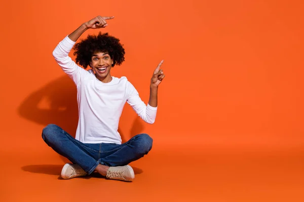 Foto de chico excitado atractivo usar camisa blanca apuntando espacio vacío sentado piso aislado color naranja fondo — Foto de Stock