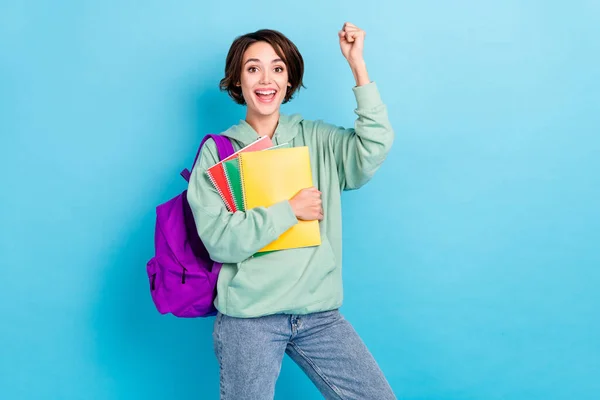 Photo of shiny lucky lady dressed green pullover backpack rising fists holding copybooks isolated blue color background — Stock Photo, Image