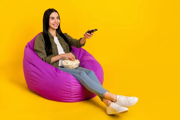 Full size photo of nice young lady look tv eat pop corn wear shirt jeans shoes isolated on yellow background