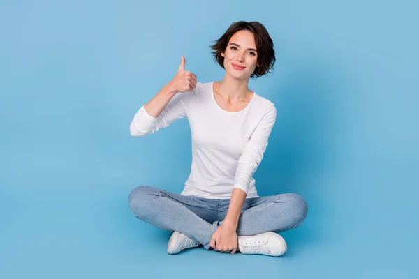 Foto de encantadora mulher bonito desgaste branco camisa sentado pernas cruzadas mostrando polegar para cima isolado azul cor de fundo — Fotografia de Stock