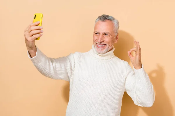 Çekici gri saçlı adamın portresi selfie çekerken bej renkli pastel arka planda izole edilmiş ok-işareti ilanı gösteriyor. — Stok fotoğraf