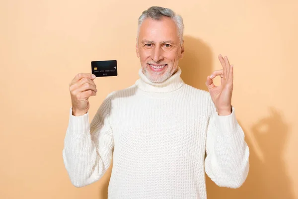 Retrato de atractivo hombre de pelo gris alegre sosteniendo en la tarjeta bancaria de la mano que muestra el anuncio ok-signo aislado sobre fondo de color pastel beige — Foto de Stock