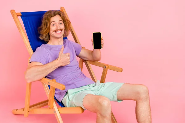 Porträtt av stilig trendig glad kille sitter i stol innehav enhet som visar tummen pekskärm isolerad över rosa pastell färg bakgrund — Stockfoto