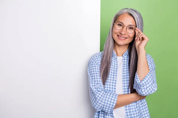 Foto van optimistisch verouderd grijs kapsel dame in de buurt van advertentie dragen bril geruite shirt geïsoleerd op groene kleur achtergrond — Stockfoto