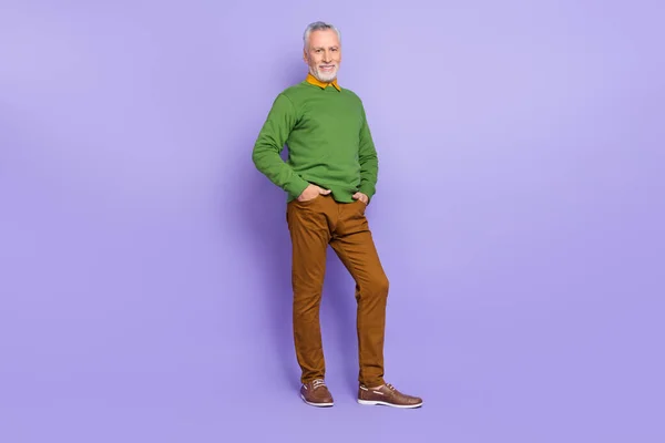Фото харизматичного пенсионера носить зеленый свитер ходьба руки карманы изолированные фиолетовый цвет фона — стоковое фото