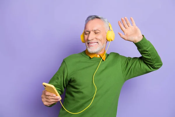 Фото веселого чоловіка на пенсії в зеленому светрі, насолоджуючись піснями сучасний пристрій танцює ізольований фіолетовий кольоровий фон — стокове фото