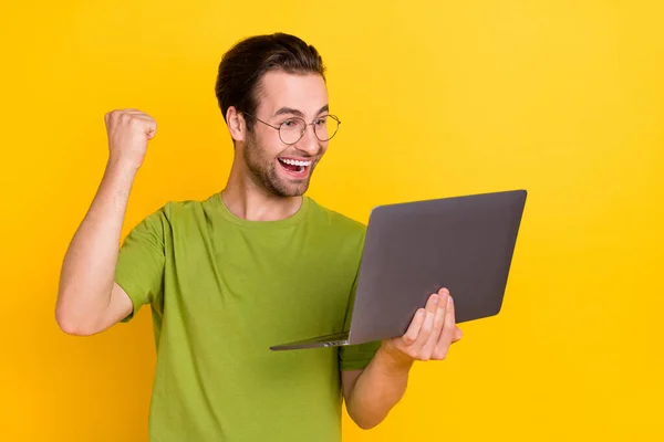 Foto van onder de indruk baard millennial guy houden laptop vuist slijtage bril groen t-shirt geïsoleerd op gele kleur achtergrond — Stockfoto