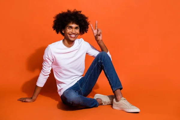 Foto di carino attraente ragazzo indossare camicia bianca che mostra v-segno seduta pavimento isolato colore arancione sfondo — Foto Stock